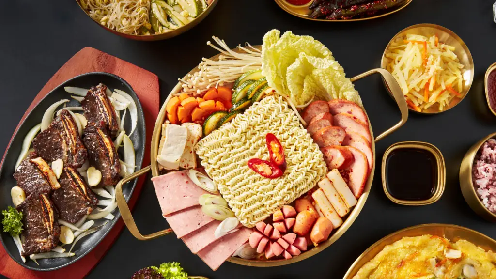 Korean budae-jjigae (army stew) with ramen noodles, spam, and vegetables in a golden hot pot, served with grilled galbi and banchan side dishes